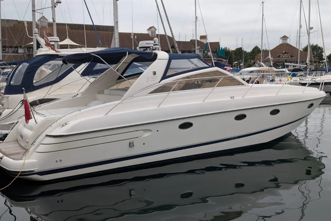 A sleek white motorboat docked in a marina, reflecting on calm water with nearby boats and buildings in the background.