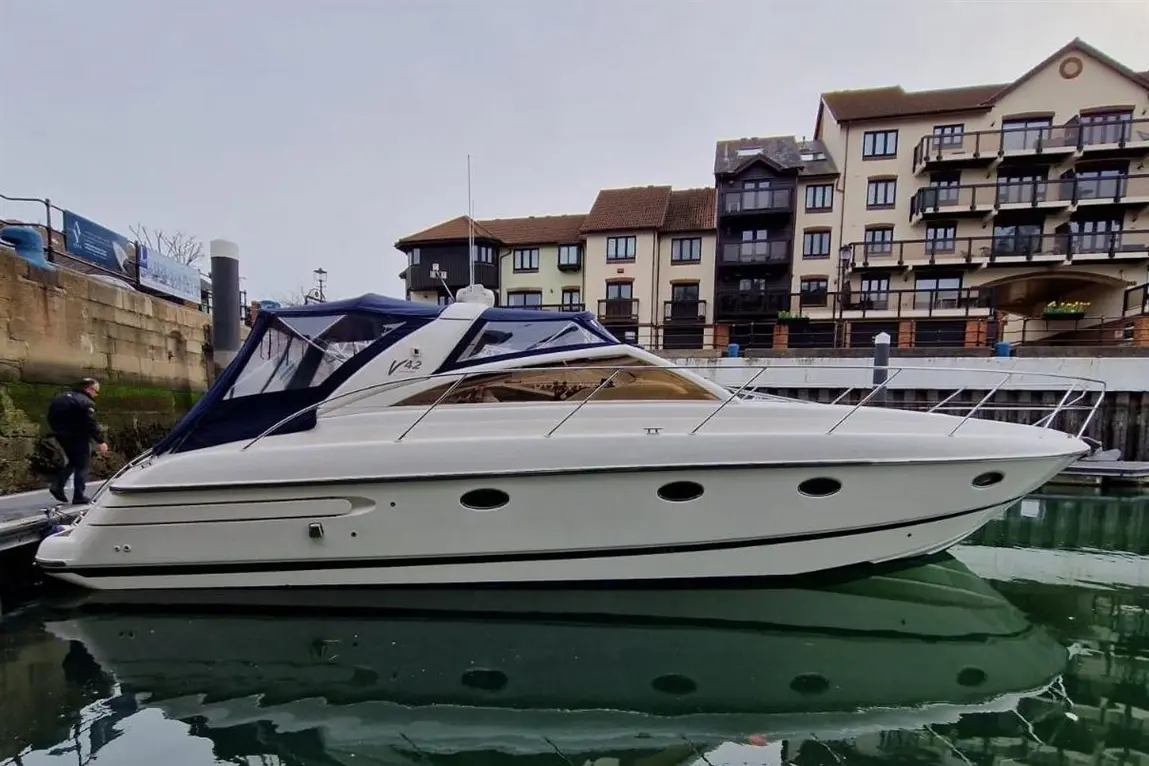 A white V42 yacht docked in a marina, with a man walking near the water and residential buildings in the background.