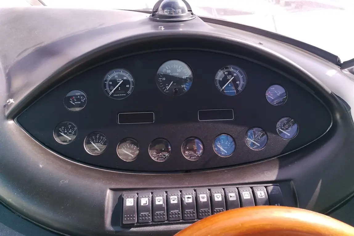 Dashboard of a boat featuring various gauges for speed, fuel, and water levels alongside control switches and a wooden steering wheel.