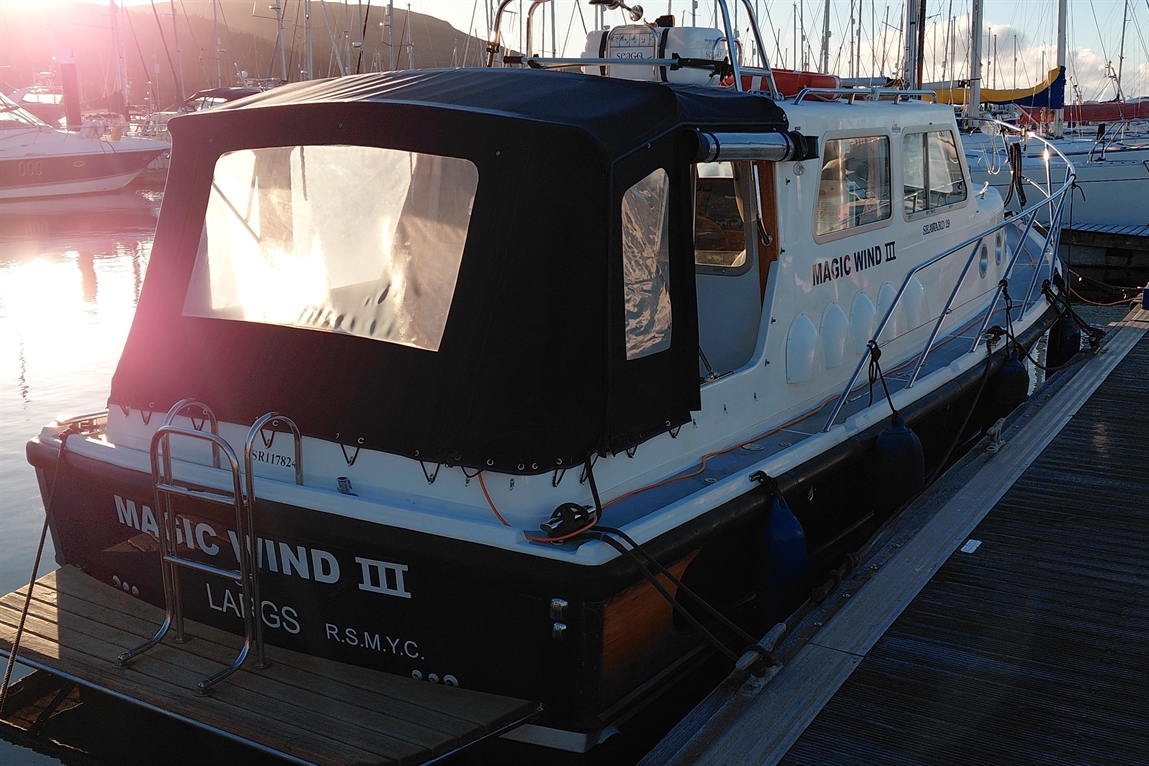 A boat named "MAGIC WIND III" is docked with a black canopy, reflecting sunlight on calm waters in a marina.
