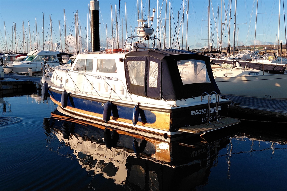 Boat named "Magic Wind III" docked at a marina, reflecting in calm water surrounded by masts of other boats.