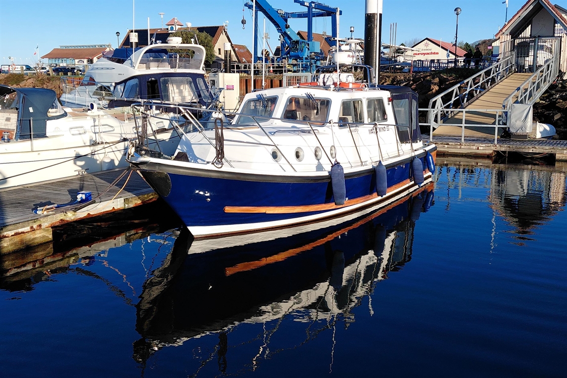 A blue and white boat is moored at a marina, reflecting on calm waters, with nearby boats and a walkway in the background.