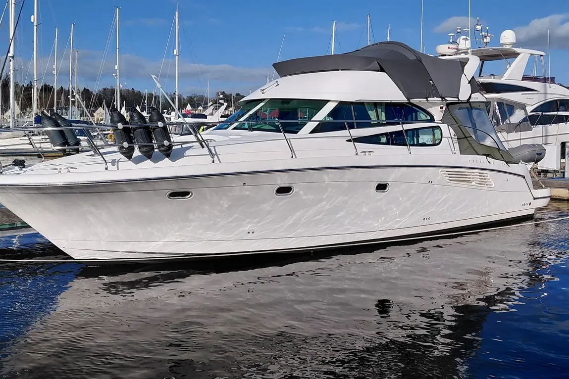 A white motor yacht docked at a marina, reflecting in the water, with several sailboats and yachts in the background.