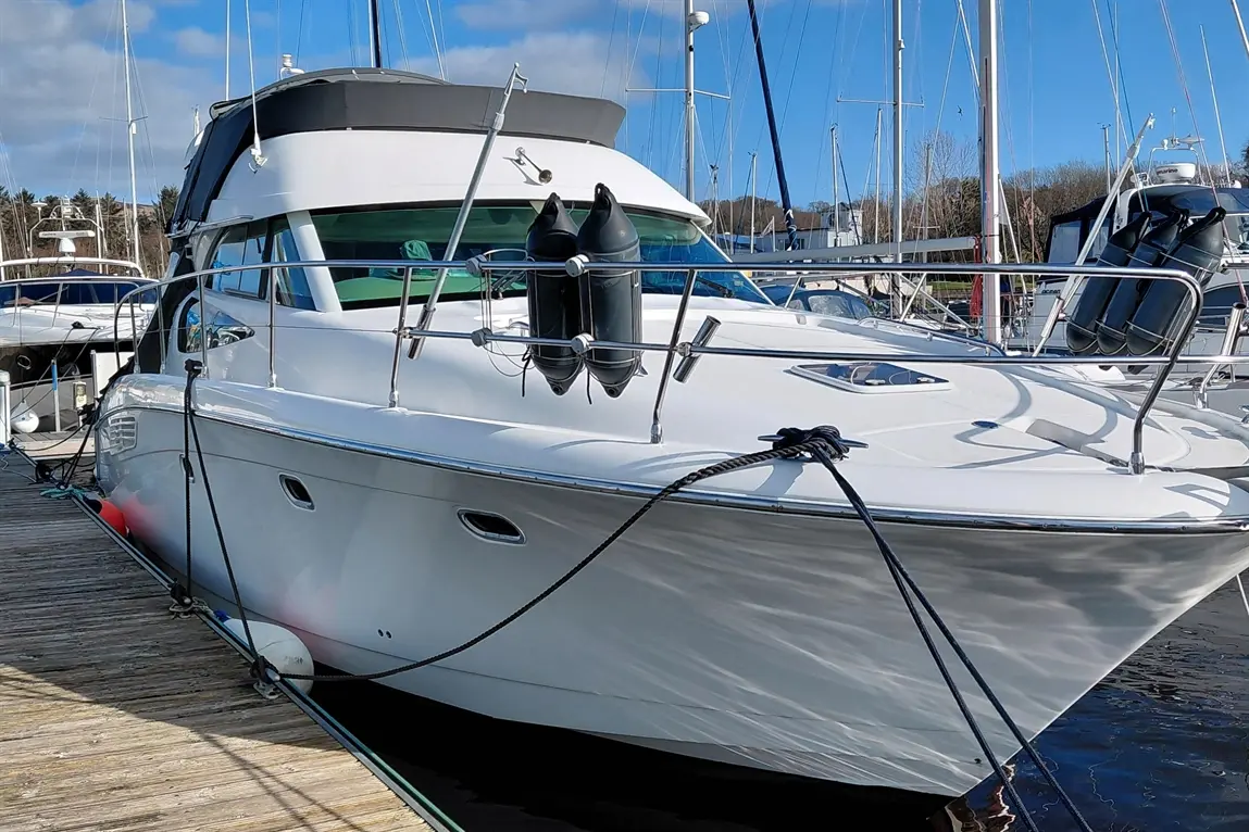 White motorboat docked at a marina, featuring fenders, a black canopy, and rigging against a blue sky with clouds.