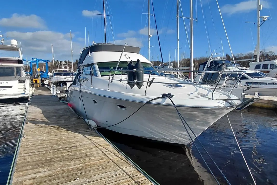 A sleek white boat moored at a marina, with wooden dock and various boats in the background under a blue sky.