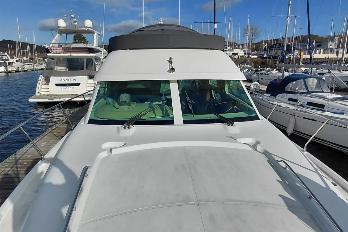 A white yacht with a grey top viewed from the front, featuring large windows and moored at a marina with other boats.