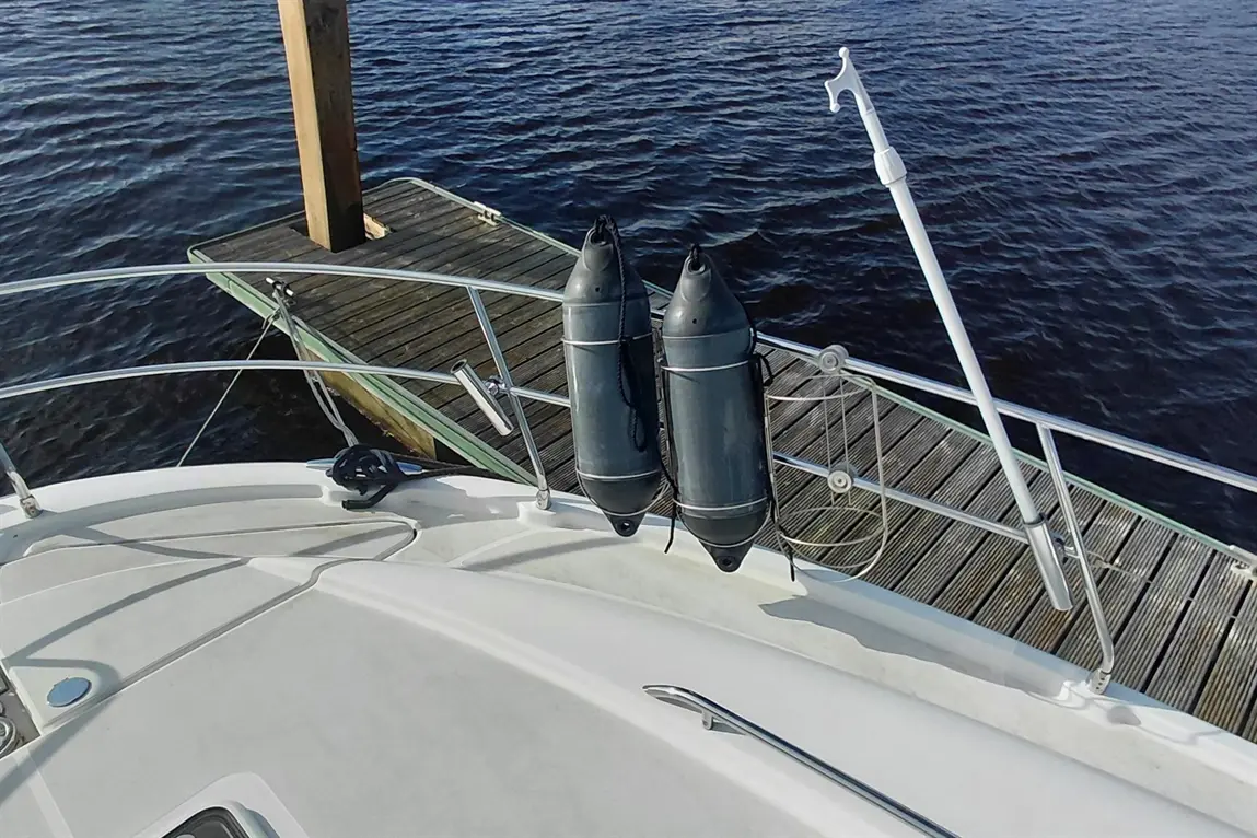 A boat deck with two black fenders secured to the railing, overlooking a wooden dock and calm water.