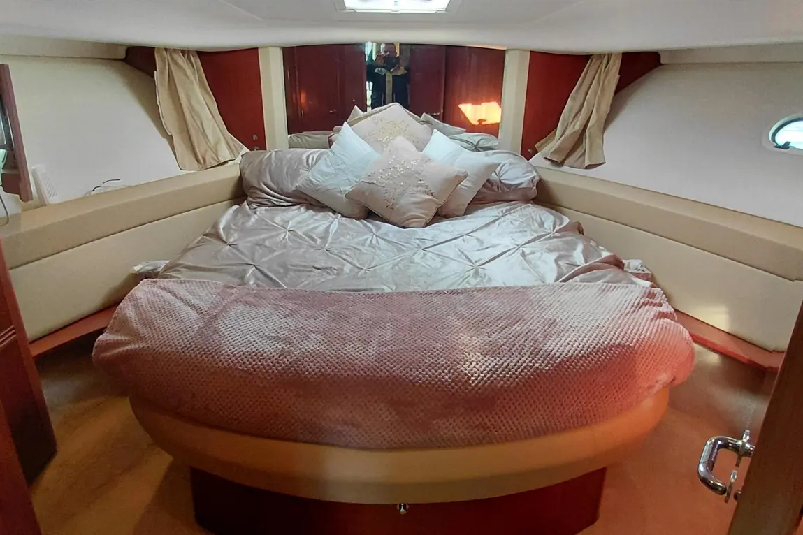 Interior of a boat cabin features a circular bed with satin bedding and decorative pillows, softly lit with a mirror reflection.
