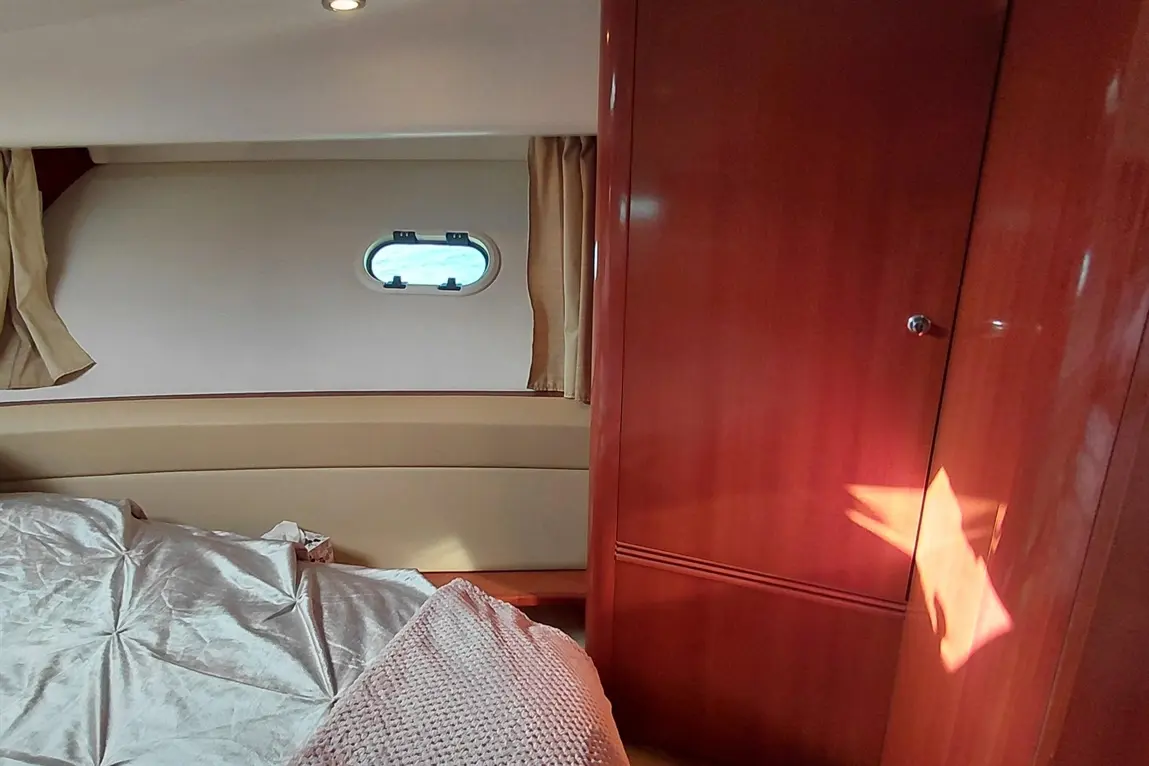 A cosy boat cabin featuring a bed with silver bedding, a wooden wardrobe, curtains, and a porthole revealing blue skies.