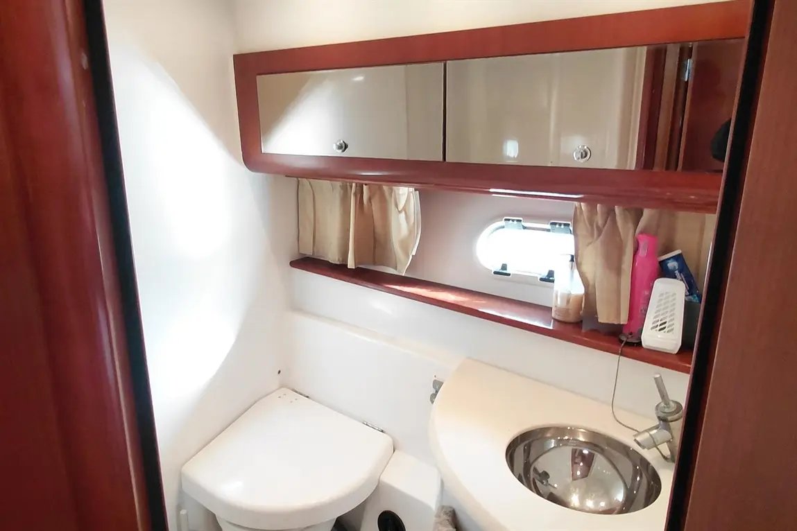 Compact marine bathroom featuring a toilet, round sink, mirror, and storage shelf with toiletries and a small window.