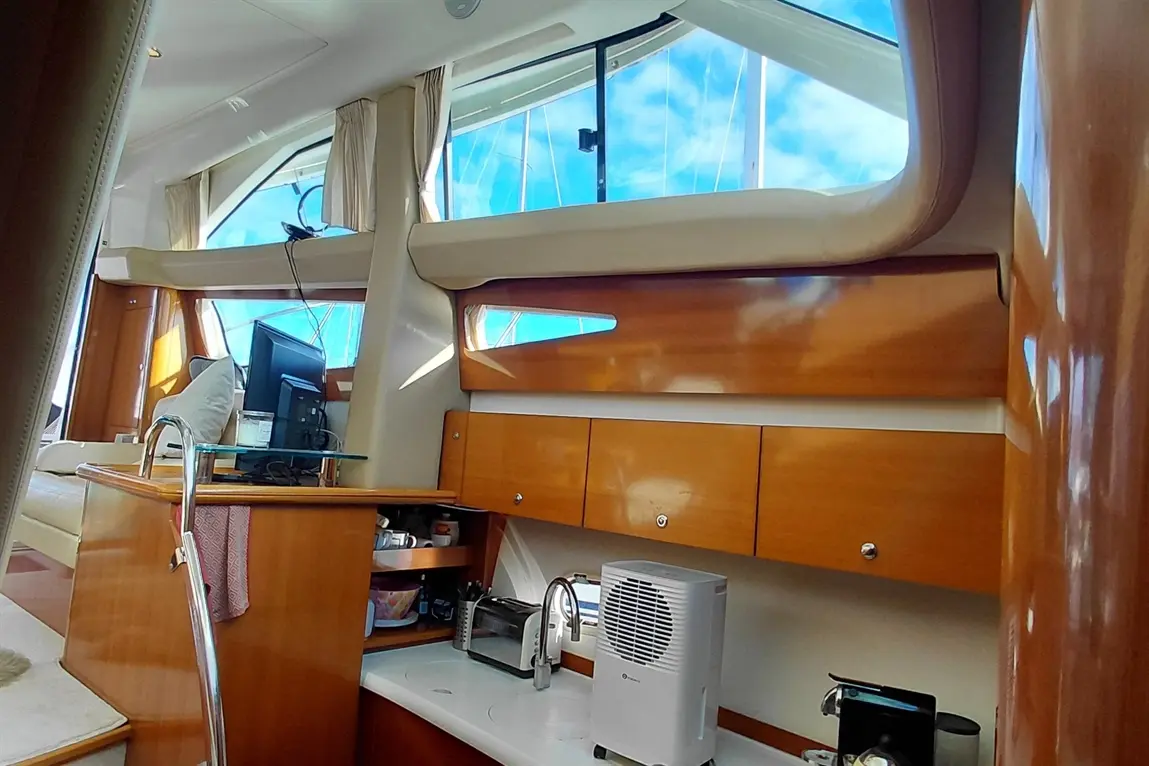 Interior of a yacht featuring a kitchenette with a computer, shelving, a toaster, coffee maker, and large windows.