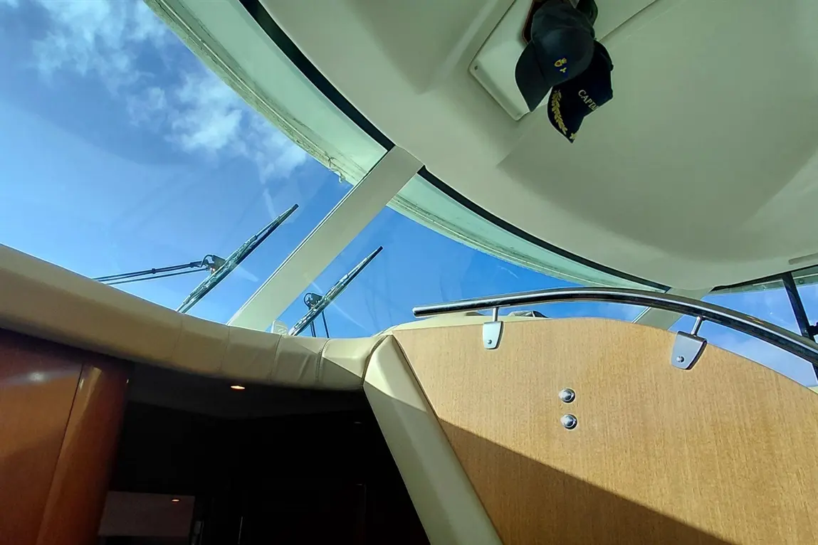 View from inside a boat, showing a clear blue sky and wipers on a curved glass window, with a captain's hat overhead.