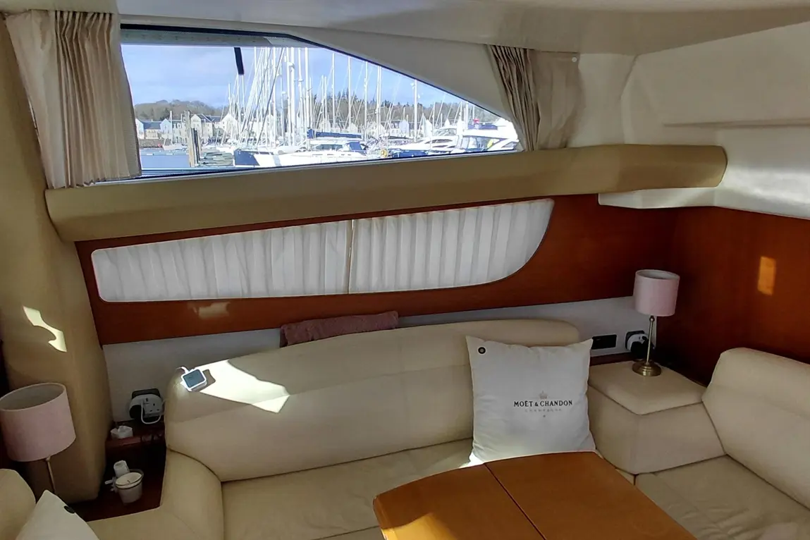 Interior of a boat cabin with cream upholstery, a wooden table, a Moët & Chandon cushion, and a view of a marina through a window.
