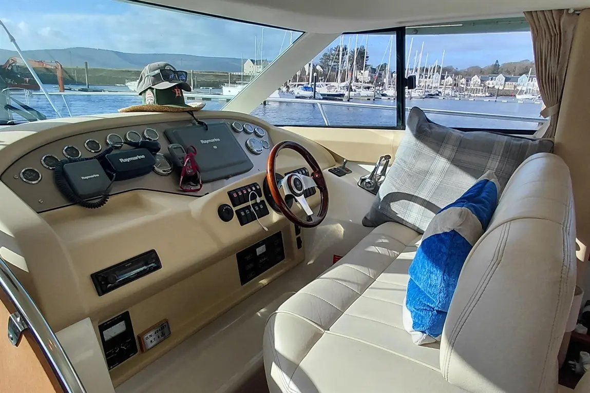 Interior of a yacht cockpit featuring steering wheel, gauges, electronics, and a view of the marina with sailboats.