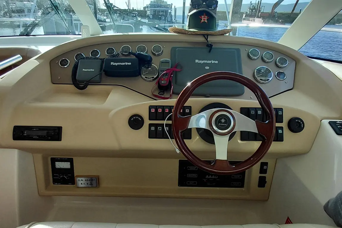 Boat cockpit with a wooden steering wheel, Raymarine equipment, buttons, and a hat displayed against a waterfront backdrop.