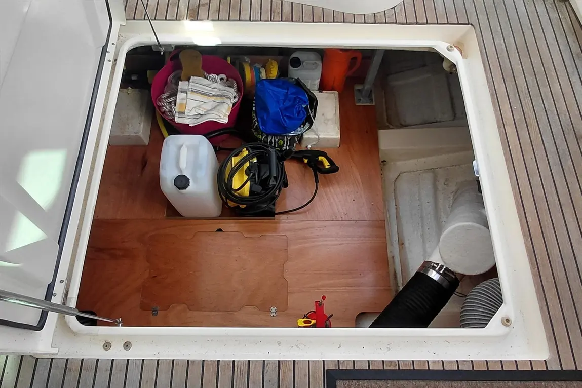 Storage hatch on a boat filled with cleaning supplies, tools, and a container, with a wooden floor visible inside.