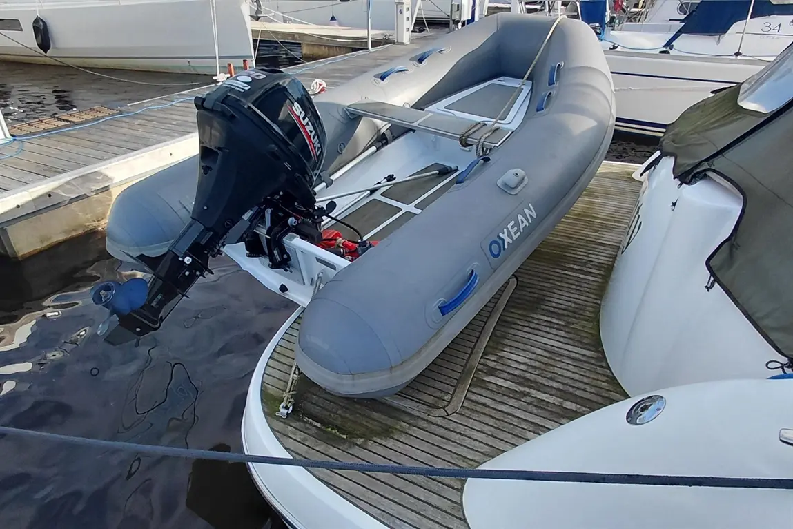 A grey inflatable dinghy with a Suzuki outboard motor is docked beside larger boats at a marina, displaying 'OXEAN' branding.