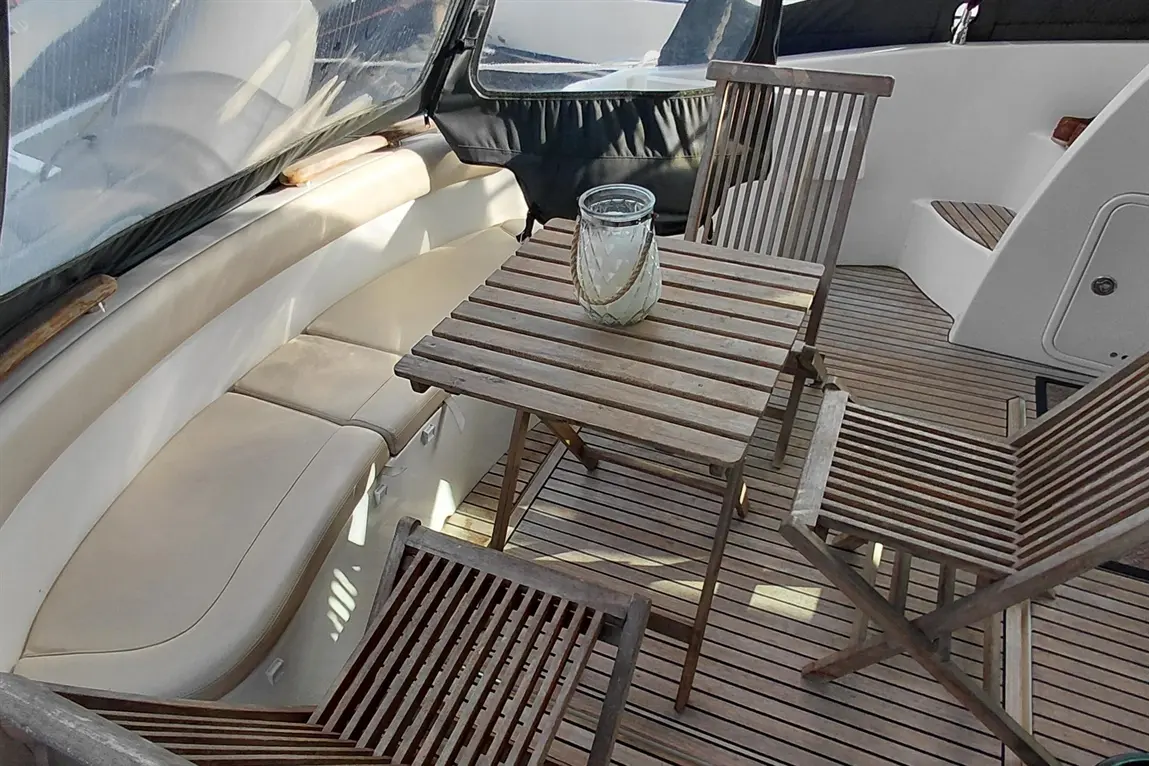 A wooden table and chairs are set on a boat's deck, with a decorative glass vase in the centre and cushioned seating nearby.