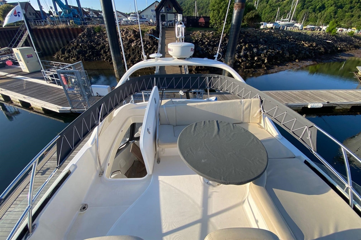 Aerial view of a yacht's deck with a circular table and white seating, docked near a marina with boats in the background.