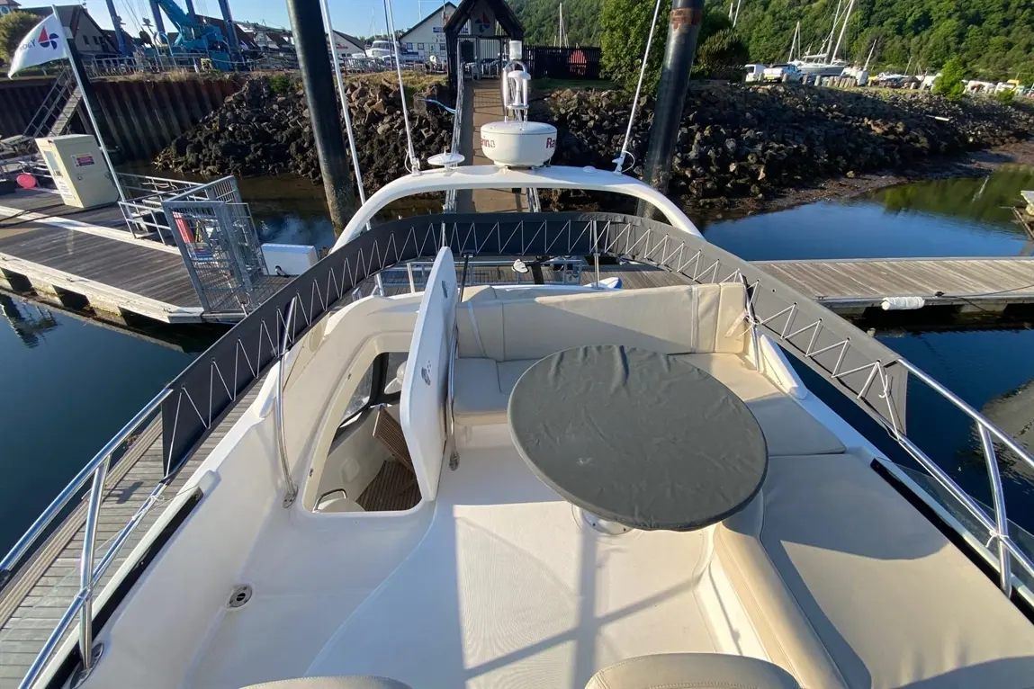 Aerial view of a yacht's deck with a circular table and white seating, docked near a marina with boats in the background.