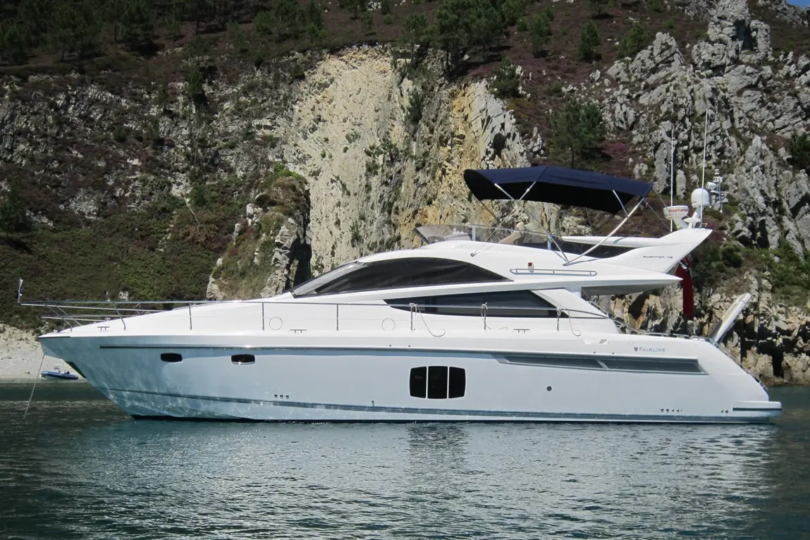 A sleek white yacht anchored near a rocky coastline with greenery, featuring a blue canopy and visible branding "Fairline.