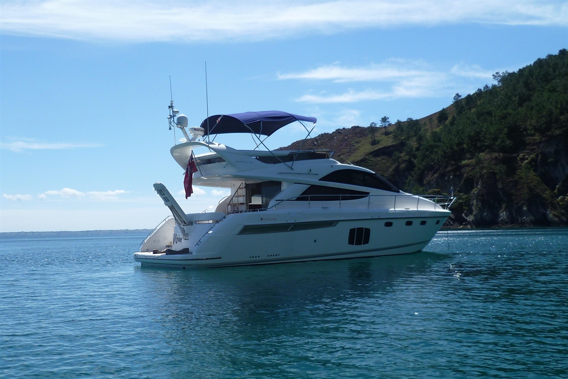 A luxurious white yacht with a dark blue canopy anchored in calm waters, surrounded by a forested shoreline under a bright sky.