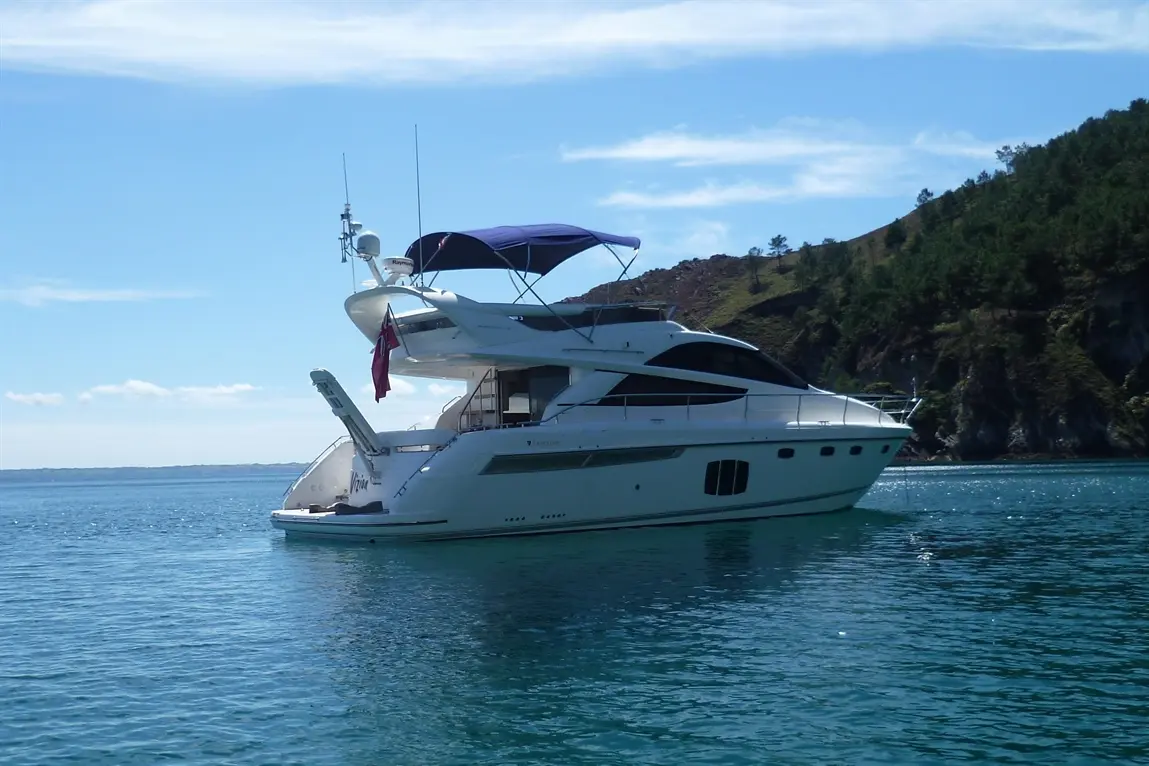 A luxurious white yacht with a dark blue canopy anchored in calm waters, surrounded by a forested shoreline under a bright sky.