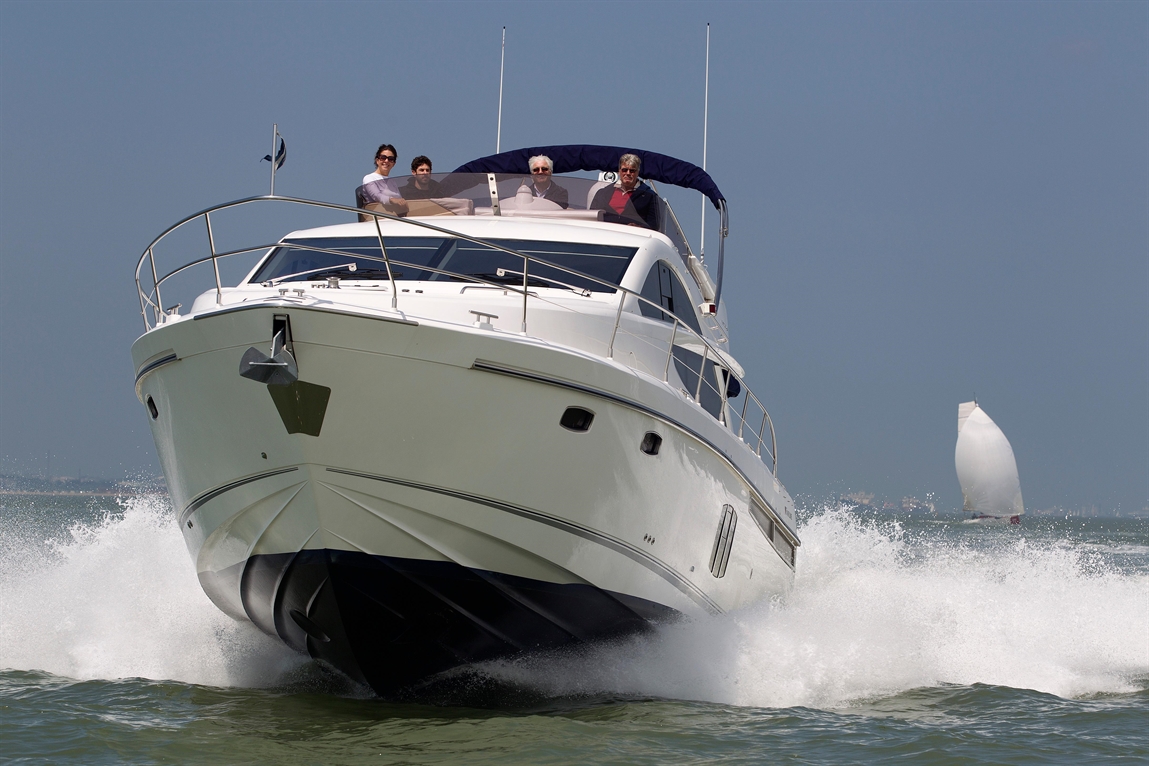 A white yacht with four people on the deck moves through the water, creating splashes, with a sailboat in the background.