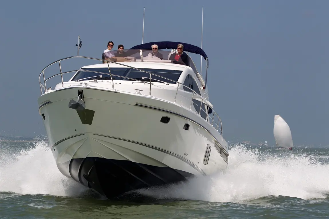A white yacht with four people on the deck moves through the water, creating splashes, with a sailboat in the background.