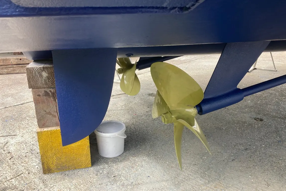 Underneath a blue boat, two yellow propellers are visible, resting on wooden supports with a white bucket nearby.
