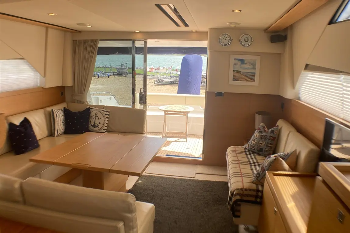 Interior of a yacht featuring a wooden dining table, plush seating, and a view of boats near a beach through large glass doors.