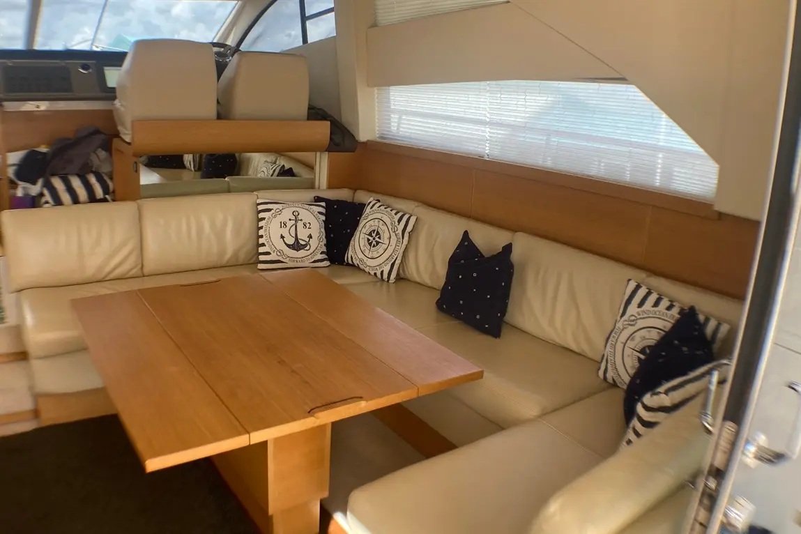 Interior of a yacht featuring a leather sofa with nautical-themed cushions and a wooden table. Windows provide natural light.
