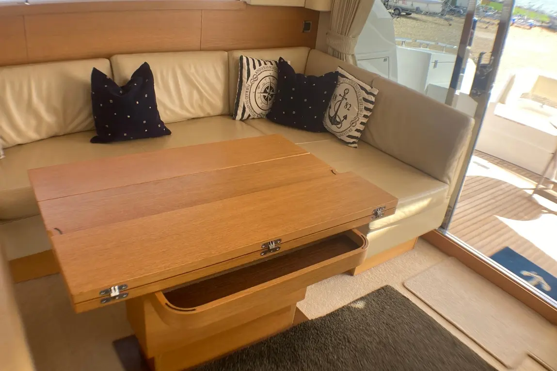 A wooden folding table in a boat's lounge beside a cream sofa with nautical-themed cushions and a view outside.