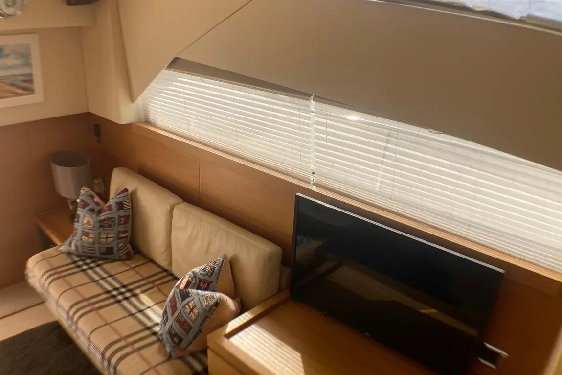 Interior of a yacht, featuring a beige sofa with patterned cushions, a lamp, and a TV, illuminated by daylight through blinds.