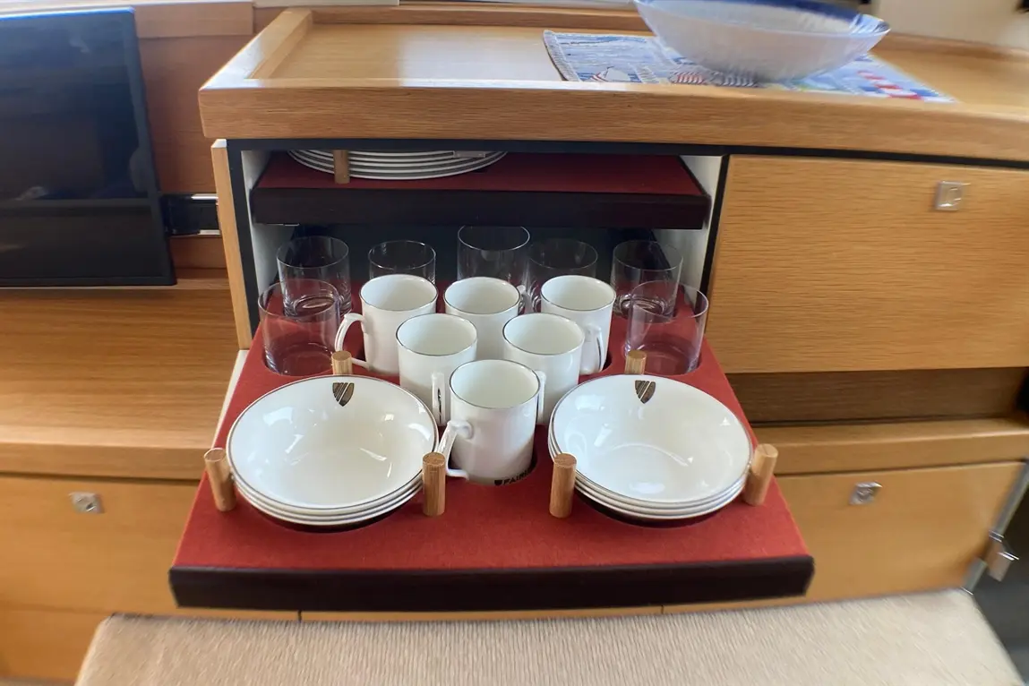 A wooden storage unit displays stacked plates, glasses, and cups on a red surface, with a bowl above and a TV nearby.