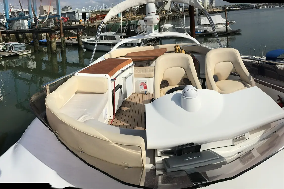 A luxury boat's deck features beige seating, a wooden table, and a helm area with marine equipment, set in a marina.