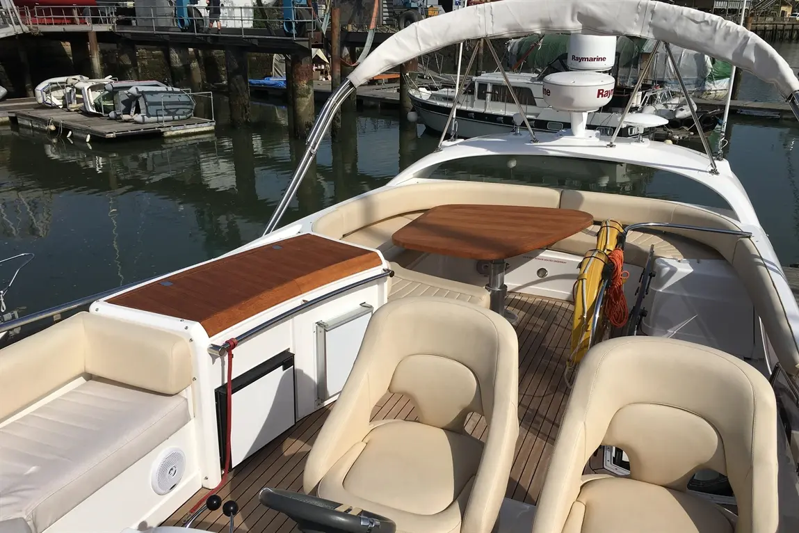 Interior of a boat featuring beige seating, a wooden table, and a cooler, with boats docked in the background.