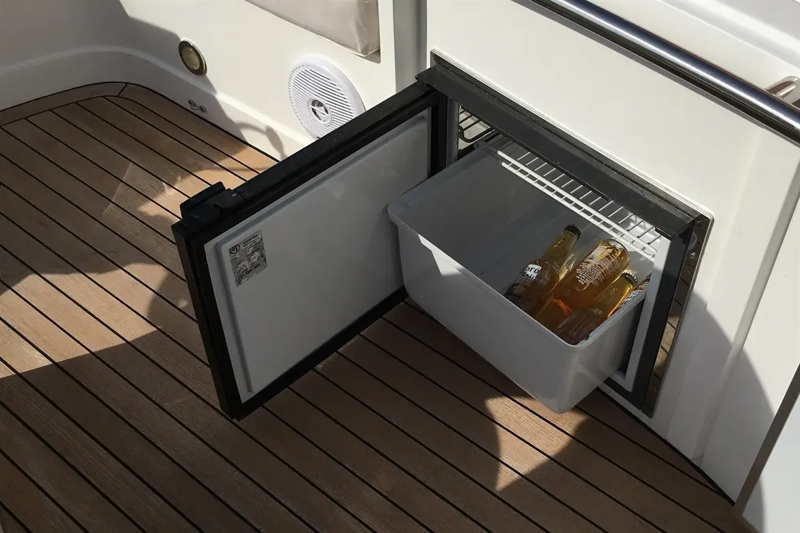 A boat's fridge with an open door, revealing a container with several bottles of Corona beer on a wooden deck.