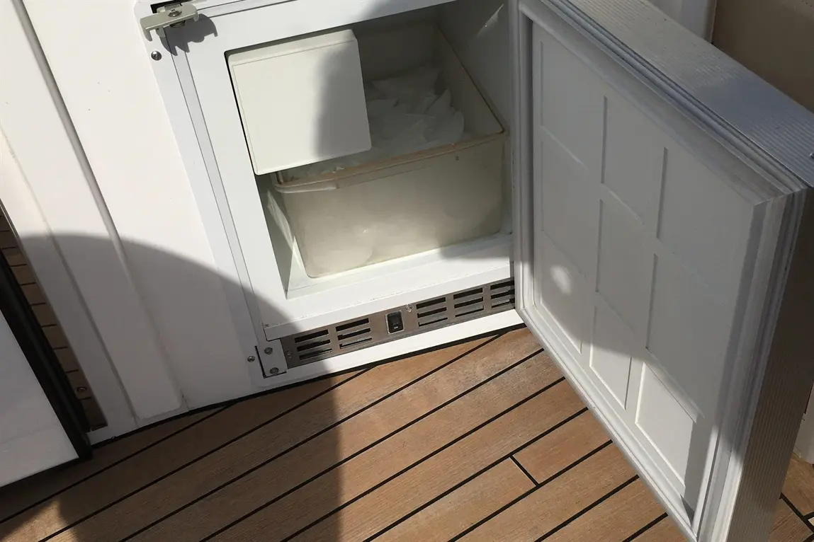A white freezer door is open, revealing a plastic container filled with ice, set on wooden decking.