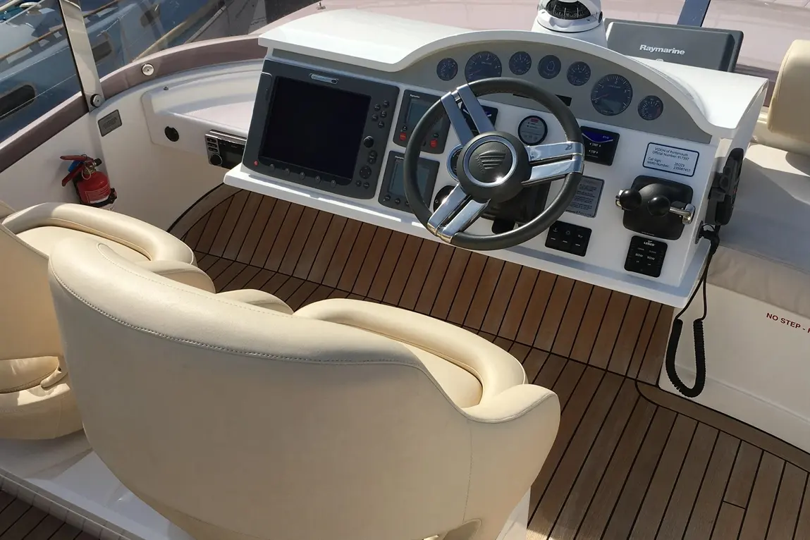 Boat cockpit with beige leather chairs, a steering wheel, and a Raymarine display surrounded by wooden flooring. Fire extinguisher present.