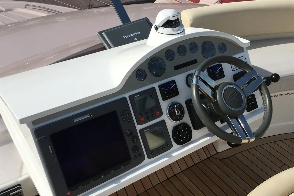 Boat control panel featuring gauges, screens, and a steering wheel on a polished deck. Mounted Raymarine display visible.