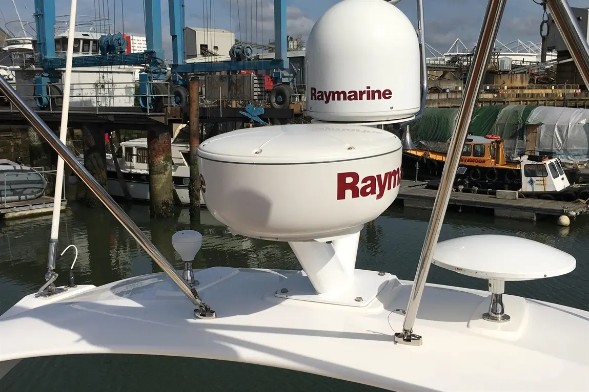 Raymarine radar and satellite device mounted on a boat, with a marina and other vessels visible in the background.