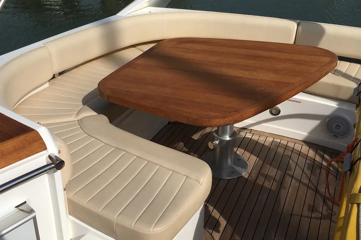 Interior of a boat featuring a round wooden table and beige cushioned seating against a backdrop of water.
