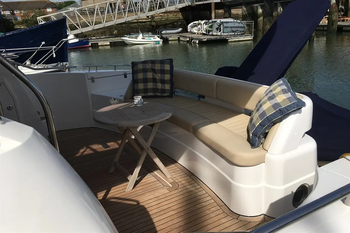 A luxury yacht's deck featuring a cushioned seating area, a wooden table, and a small cup, with boats in the marina.