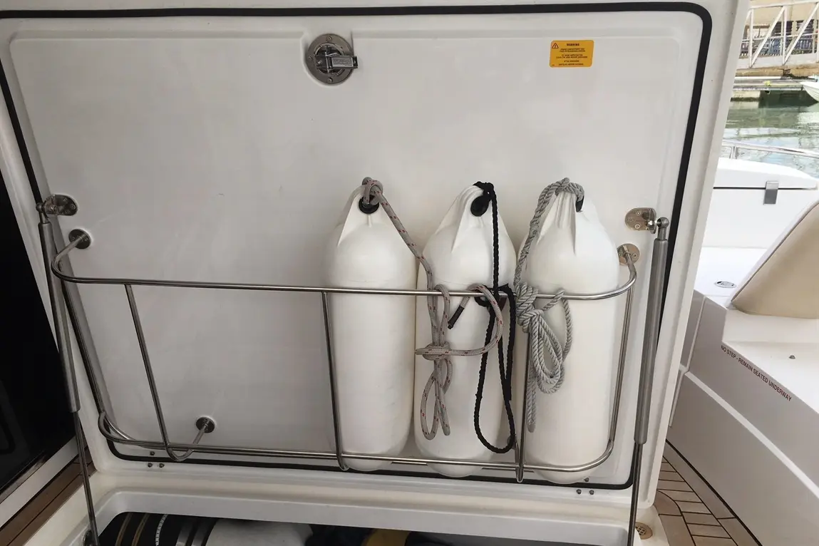 Three white fenders secured in a stainless steel holder on a boat, with a warning label on the door behind them.