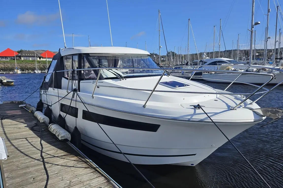 A modern white motorboat is docked at a marina, moored with ropes, surrounded by masts and water under a clear blue sky.