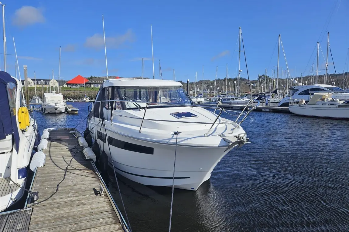 White motorboat docked in a marina, with other boats and a blue sky in the background, reflecting calm water.