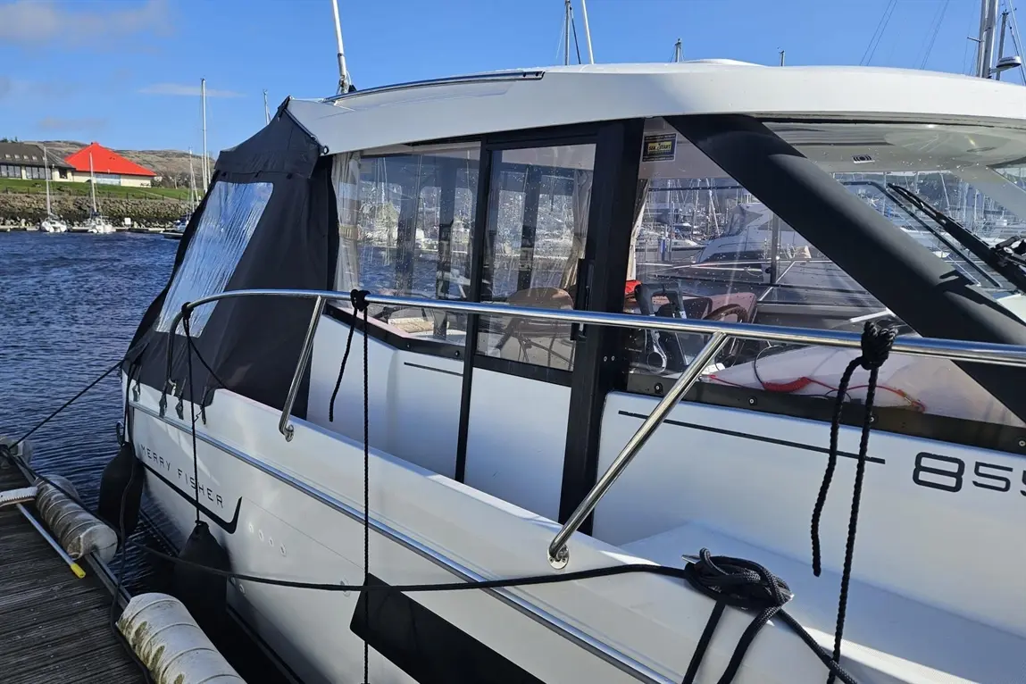 A modern boat named 'Merry Fisher' docked beside a marina, with clear blue water and a red-roofed building in the background.