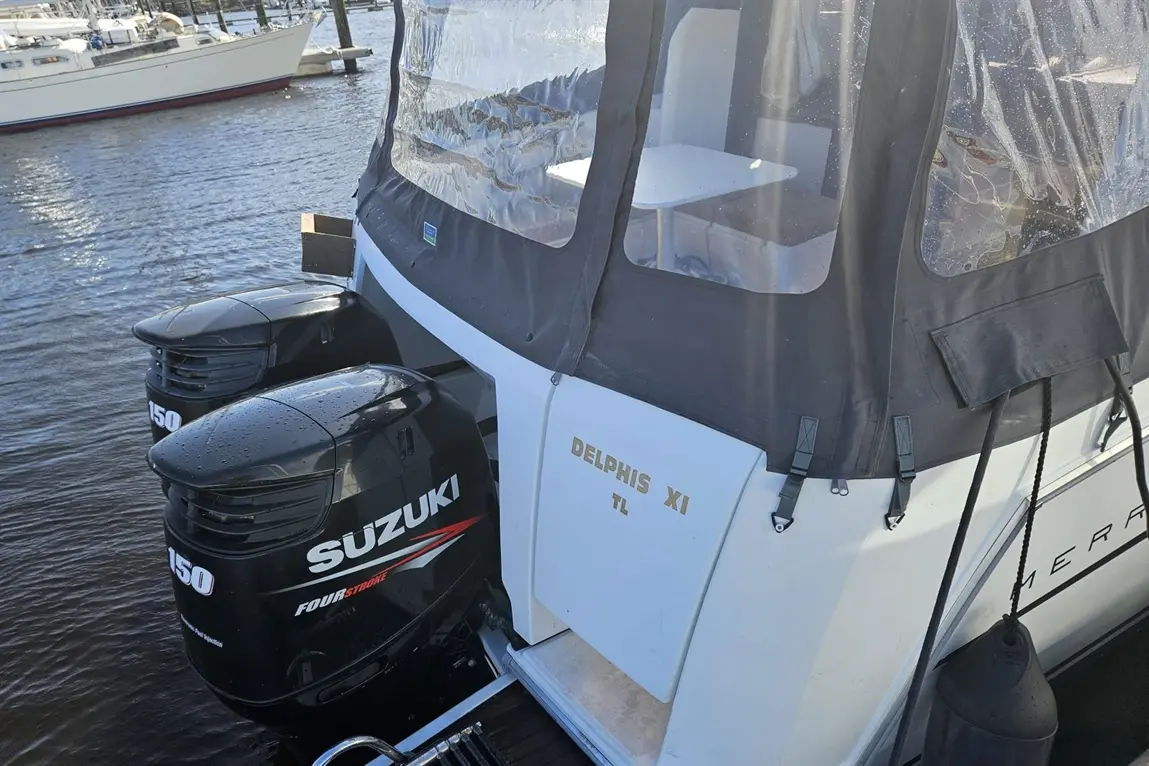Two Suzuki outboard motors on a boat named "Delphis XI" docked by a marina, with clear water and other boats in the background.