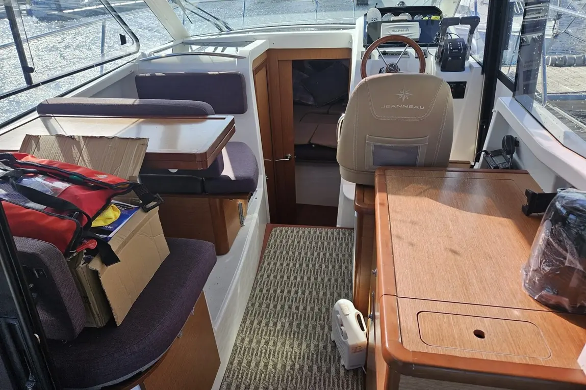 Interior of a Jeanneau boat, featuring seating, a steering wheel, navigation equipment, and a doorway leading to the cabin.
